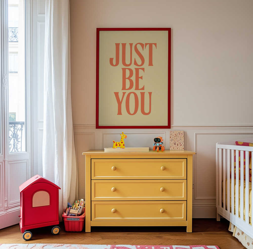 Framed nursery poster with 'JUST BE YOU' text in orange on a light yellow background hanging on the wall in the children's room